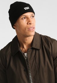 Beanie nero a maglia con un piccolo etichetta bianca del logo Levi's. Indossato con un giubbotto con colletto marrone sopra una maglietta a righe, su uno sfondo chiaro.