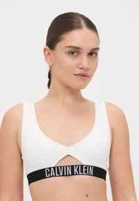 Bralette blanc à côtes avec une découpe en forme de V et une ceinture noire sur laquelle est inscrit "CALVIN KLEIN" en texte blanc. Texture lisse, design minimal.
