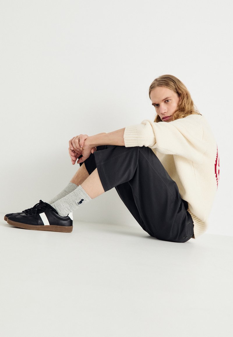 Jersey de punto crema, pantalones cortos grises de pierna ancha, calcetines grises acanalados y zapatillas negras con rayas blancas. Pose sentada, fondo neutro.