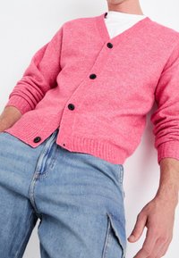 Różowy sweter kardigan z dekoltem w serek i czarnymi guzikami. Posiada prążkowane mankiety i dół oraz fakturowaną tkaninę. Połączony z jasnoniebieskimi dżinsami.