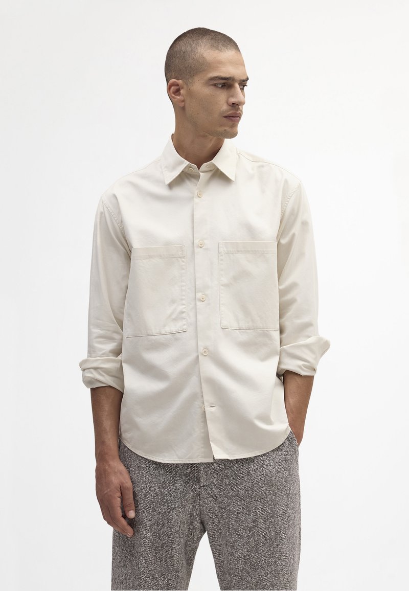 Lys beige skjorte med knapper og to brystlommer, lange ærmer opkrøllede ved albuerne, lavet af et glat stof.