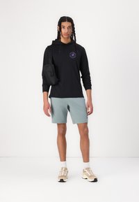 Hoodie noir à manches longues avec logo, shorts bleu clair et baskets beiges avec des chaussettes blanches texturées. Sac bandoulière noir sur l'épaule.