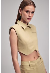 Geknipt mouwloos shirt in lichtbeige materiaal, met een puntige kraag en een voorzakdetail. Gladde textuur met een gestructureerd ontwerp.