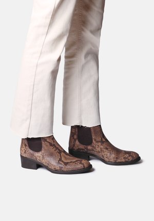 Bottines en cuir imitation serpent marron avec des panneaux élastiques sur les côtés et un talon carré modeste, associées à un pantalon crème avec un ourlet effiloché.