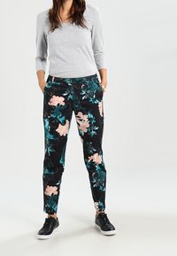 Schwarze Blumenhose mit rosa und Türkis floralem Muster, aus weichem Material gefertigt. Kombiniert mit einem grauen Langarmoberteil und schwarzen Turnschuhen.