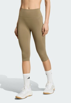 Kvinde iført beige cropped leggings med høj talje og sidelomme, hvide crew-sokker og hvide sportssko, stående mod en ensfarvet baggrund.