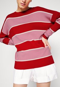 Pull rayé rouge et rose à coupe ample, avec poignets côtelés et encolure arrondie. Associé à un short blanc.