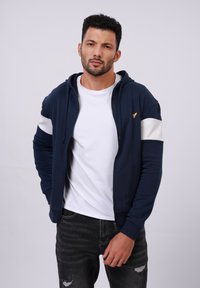 Hoodie zippé marine avec des accents de rayures blanches et un logo sur la poitrine. Tissu mélangé coton, offrant une coupe décontractée et une capuche réglable à cordon.