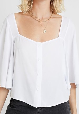 Blouse blanche boutonnée avec encolure carrée et manches courtes amples, associée à des colliers en chaînes dorées superposées et un pantalon sombre.