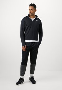 Mężczyzna w czarno-szarym dresie sportowym, w białych skarpetach i czarnych sneakersach, stojący na tle jednolitego białego tła.