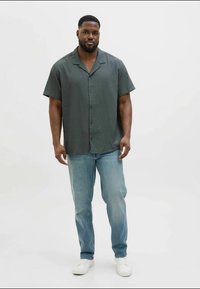 Camicetta a maniche corte verde scuro con bottoni e motivo testurizzato, abbinata a jeans azzurri chiari e sneakers bianche.