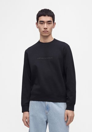 Junger Mann mit kurzen schwarzen Haaren trägt einen schwarzen Emporio Armani Sweatshirt und hellblaue Jeans vor weißem Hintergrund.