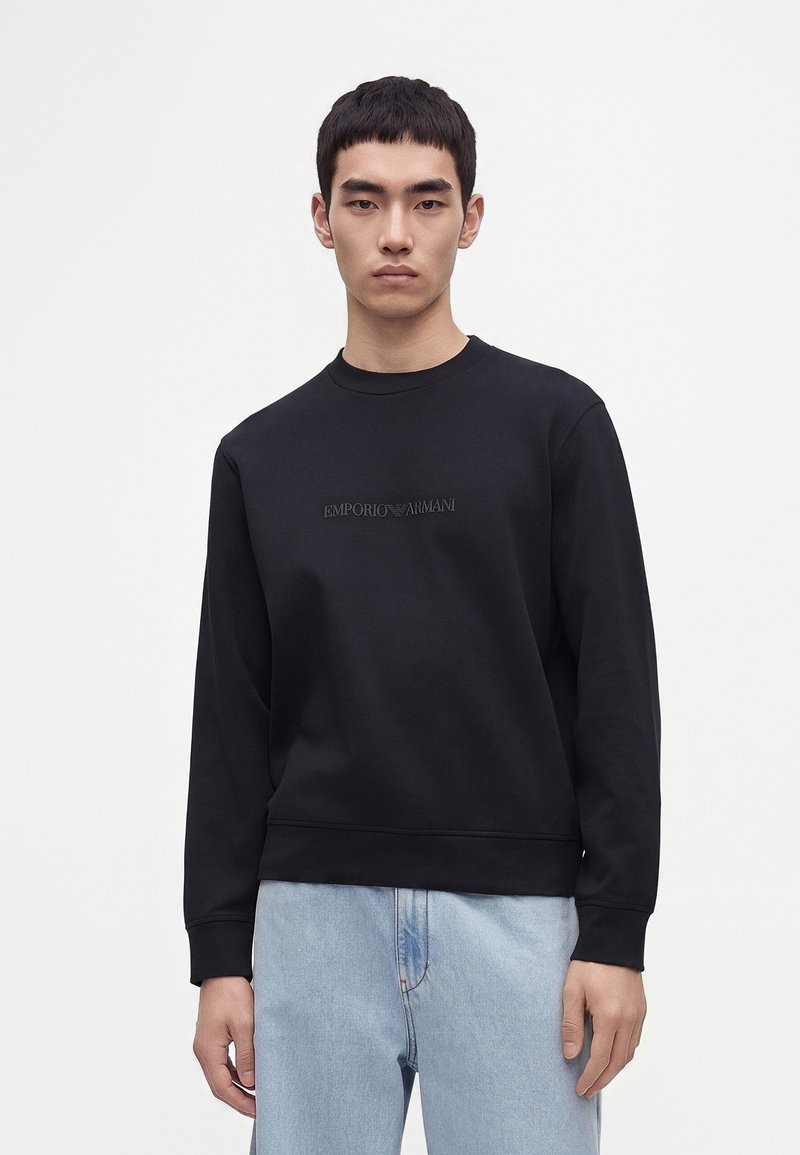 Jeune homme aux cheveux courts et noirs porte un sweat-shirt noir Emporio Armani et un jean bleu clair sur un fond blanc uni.