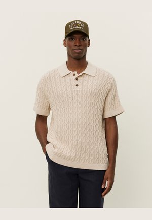 Beige Strick-Polohemd mit strukturiertem Muster, kurzen Ärmeln und einer Knopfleiste mit zwei Knöpfen. Kombiniert mit einer dunklen Mütze mit weißem Text.