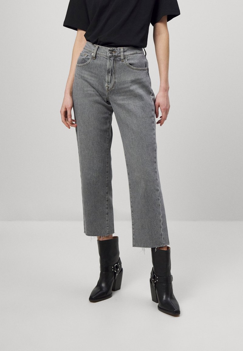 Person in verkürzten grauen Jeans, schwarzem Shirt und schwarzen, mit Schnallen verzierten, hohen Stiefeletten vor hellem Hintergrund.