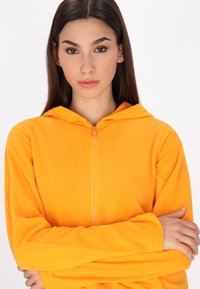 Felpa arancione con zip e cappuccio. Tessuto morbido e maniche lunghe, con una zip centrale e vestibilità rilassata.
