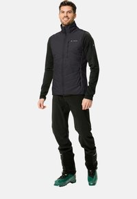 Gilet isolato nero con colletto alto, tasche con zip, indossato sopra una maglietta nera a maniche lunghe e pantaloni neri, completato da stivali da sci verdi.