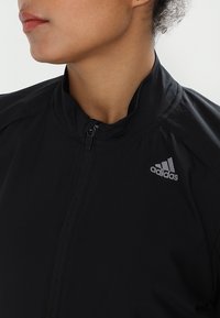 Veste de sport noire avec un col haut, fermeture éclair et logo Adidas gris sur la poitrine. Texture de tissu lisse.