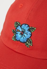 Raudona medvilninė beisbolo kepurė su mėlynu siuvinėtiniu hibisko žiedu, aplink kurį yra žali lapai ir juodos kontūro linijos priekyje.