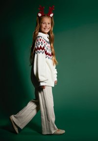 Kremowy sweter typu pulower z czerwono-niebieskim geometrycznym wzorem, w połączeniu z jasnymi, szerokimi spodniami o fakturze oraz beżowymi butami. Czerwona opaska na głowę z reniferem.