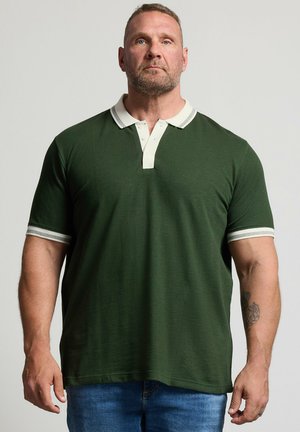 Muskulöser Mann mit kurzen Haaren und Bart, der ein dunkelgrünes Poloshirt mit weißem Kragen und blaue Jeans trägt, steht vor einfarbigem Hintergrund.