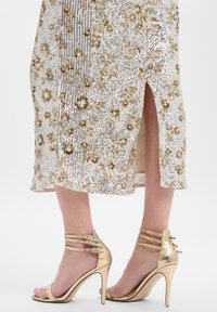 Goldene Sandalen mit hohem Absatz und metallischem Finish, mit Knöchelriemen und Peek-toe-Design, kombiniert mit einem paillettenbesetzten Kleid mit Blumenmuster.