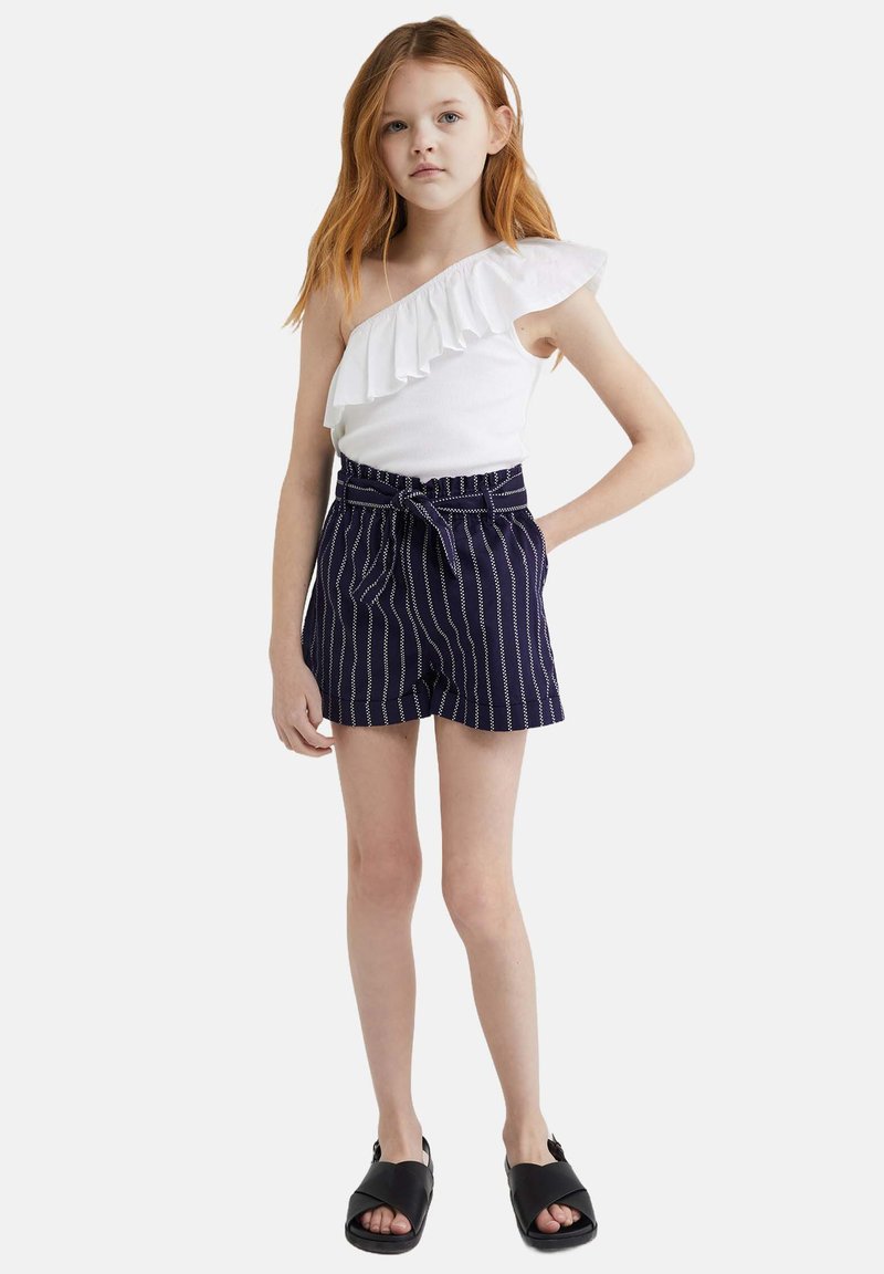 Balta vienas pleca blūze ar volāna akcentu, saskaņota ar jūru svītrainām šortiem, kas papildināti ar auklu. Melni sandales noslēdz apģērbu.