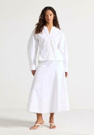 Kvinde iført hvid bluse med lange ærmer og matchende midi-nederdel med små blomsterbroderier, sammen med beige sandaler med hæl, stående afslappet.