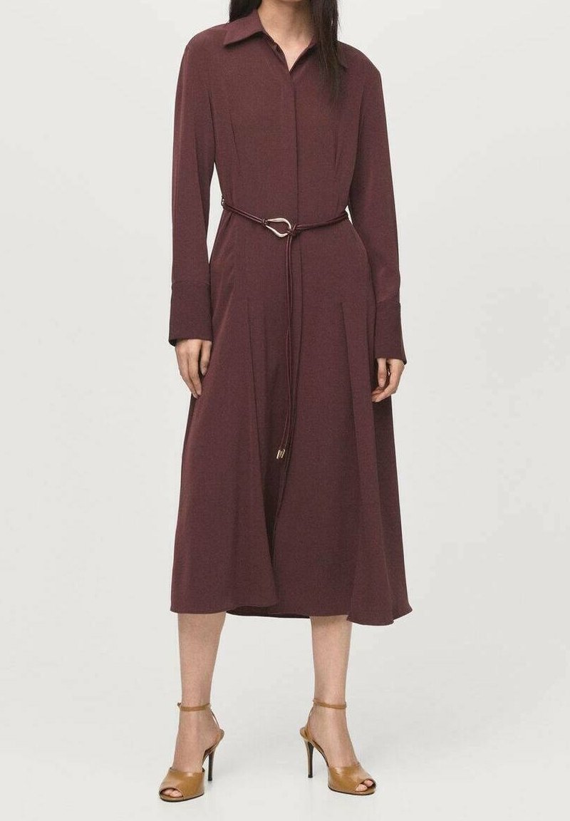 Femme portant une robe midi bordeaux à manches longues et col, avec une fine ceinture et des sandales à talons beiges sur un fond uni.