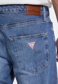 Vaqueros de mezclilla azul con un bolsillo trasero que presenta un parche triangular de logo de Guess en rojo y blanco. Etiqueta de cuero marrón en la cinturilla con marcas de la marca.