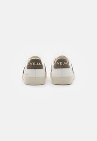 White sneakers with a beige suede heel tab, featuring 'VEJA' branding in white on the heel. The design has a minimalist style.