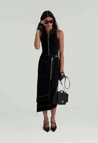 Black sleeveless dress with a zip front, side pockets, and asymmetrical hem. Accented with white stitching. Paired with black heels and handbag.