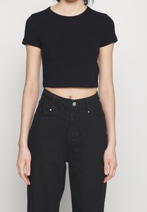 Donna che indossa un top corto a maniche corte nero e jeans neri a vita alta su uno sfondo neutro.