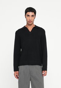 Maglione nero a collo V con colletto, orlo e polsini a coste, realizzato in un materiale morbido. Indossato sopra pantaloni grigi.