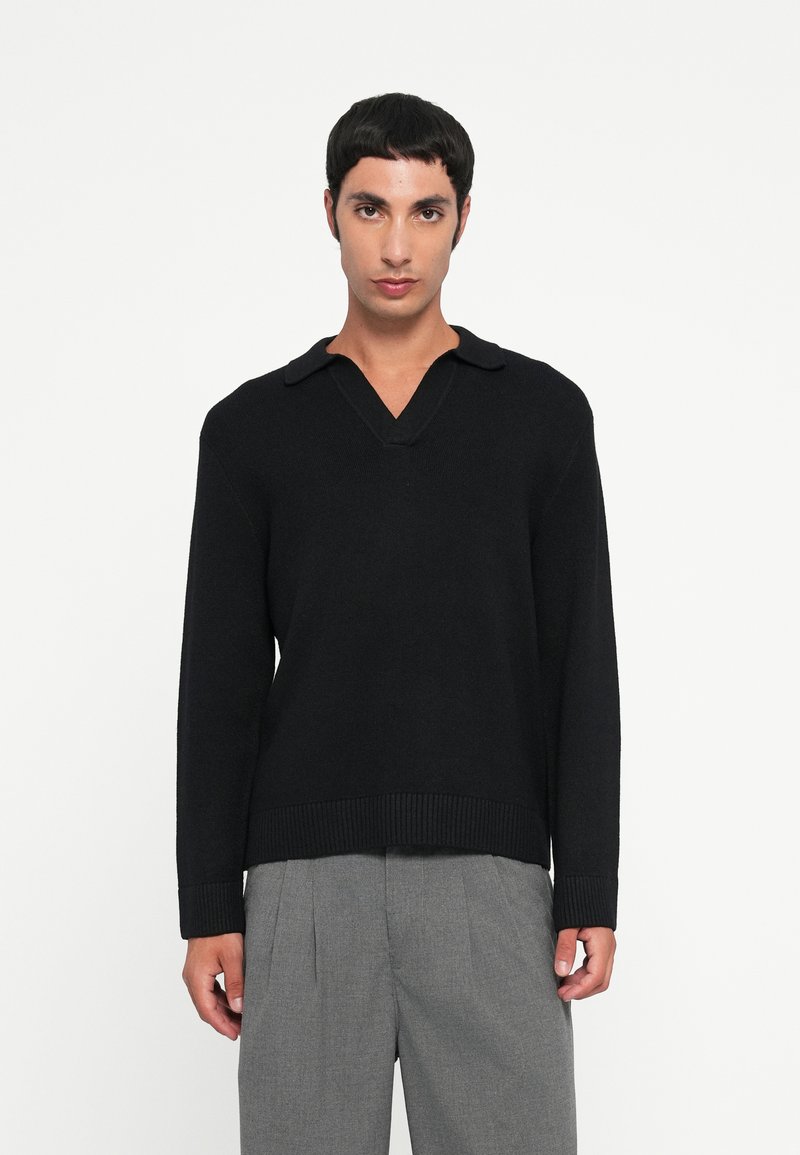 Maglione nero a collo V con colletto, orlo e polsini a coste, realizzato in un materiale morbido. Indossato sopra pantaloni grigi.