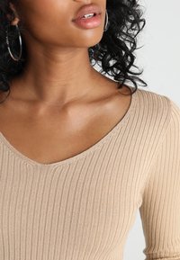 Krémový hnací pletený top s hlubokým V-výstřihem, přiléhavým střihem, dlouhými rukávy a jemnou texturou, zdůrazňující hladký finiš.