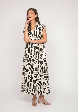 Femme souriante, portant une robe maxi sans manches à boutons avec des motifs abstraits noirs et des sandales à talons beiges, debout devant un fond uni.