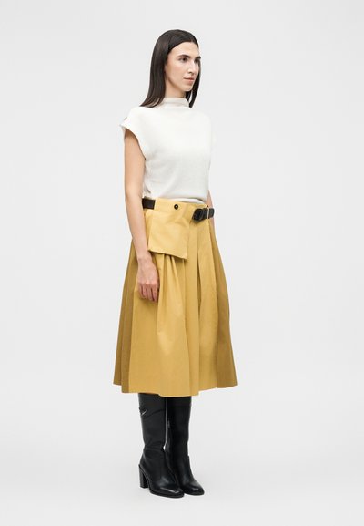 Γυναίκα με μακριά σκούρα μαλλιά που φοράει λευκό αμάνικο τοπ, μουσταρδί πλισέ μίντι φούστα με ζώνη-τσάντα και μαύρα ψηλοτάκουνα μπότες μέχρι το γόνατο.