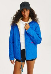 Modrá prošívaná bunda s béžovým límcem, zapínáním na zip a bočními kapsami. Obsahuje růžový akcent vpředu a texturovaný povrch.