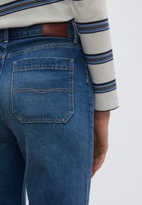 Τζιν τζιν σε μεσαία μπλε απόχρωση, με πίσω τσέπη που έχει καμπυλωτή ραφή. Η μέση φέρει καφέ δερμάτινο ταμπελάκι με λογότυπο.