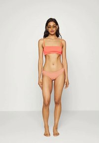 Mulher vestindo um biquíni cor-de-rosa e óculos de sol, em pé descalça contra um fundo cinza claro liso, voltada para frente com os braços relaxados.