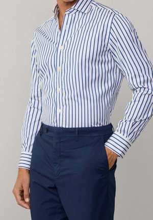 Homme portant une chemise blanche à rayures bleues verticales et un pantalon bleu marine, une main dans la poche, sur un fond clair uni.