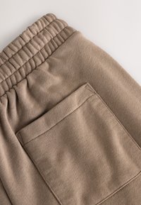 Gros plan d'un pantalon en tissu beige montrant une ceinture élastique et une poche arrière rectangulaire avec des coutures visibles.
