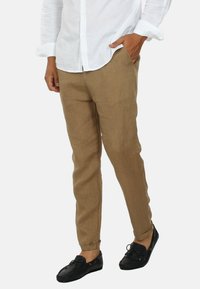Pantaloni in lino marrone chiaro con una vestibilità rilassata, dotati di tasche laterali e orli arrotolati, abbinati a scarpe nere slip-on.