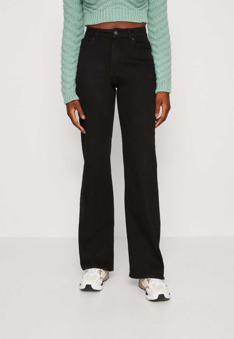 Femme portant un jean noir taille haute à jambes larges, un pull court vert clair en maille et des baskets blanches, debout devant un fond blanc.