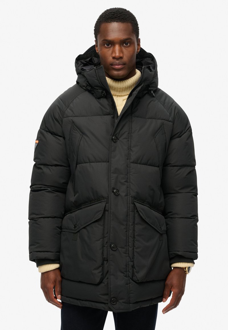 Sort polstret parka med hætte, der har to frontlommer, knaplukning og en struktureret overflade. Bæres over en beige turtleneck-sweater.
