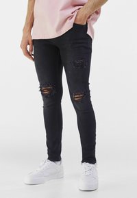 Muž v černých roztrhaných skinny džínách, bílých teniskách a světlém růžovém tričku, s jednou rukou v kapse.