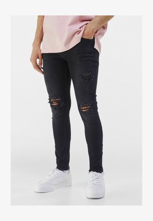 Hombre vistiendo jeans skinny rasgados negros, zapatillas blancas y una camisa rosa clara con una mano en el bolsillo.