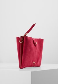 Punainen mokkahandbag, jossa on jämäkkä muoto, varustettuna yläkahvalla, kultaisilla yksityiskohdilla ja napsauskiinnityksellä, sekä logosomiste edessä.