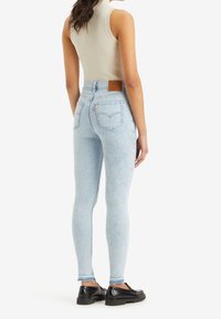 Vaqueros ajustados de cintura alta en azul claro, confeccionados en denim, con un parche de cuero en la cinturilla y dos bolsillos traseros. Se llevan zapatos gruesos.